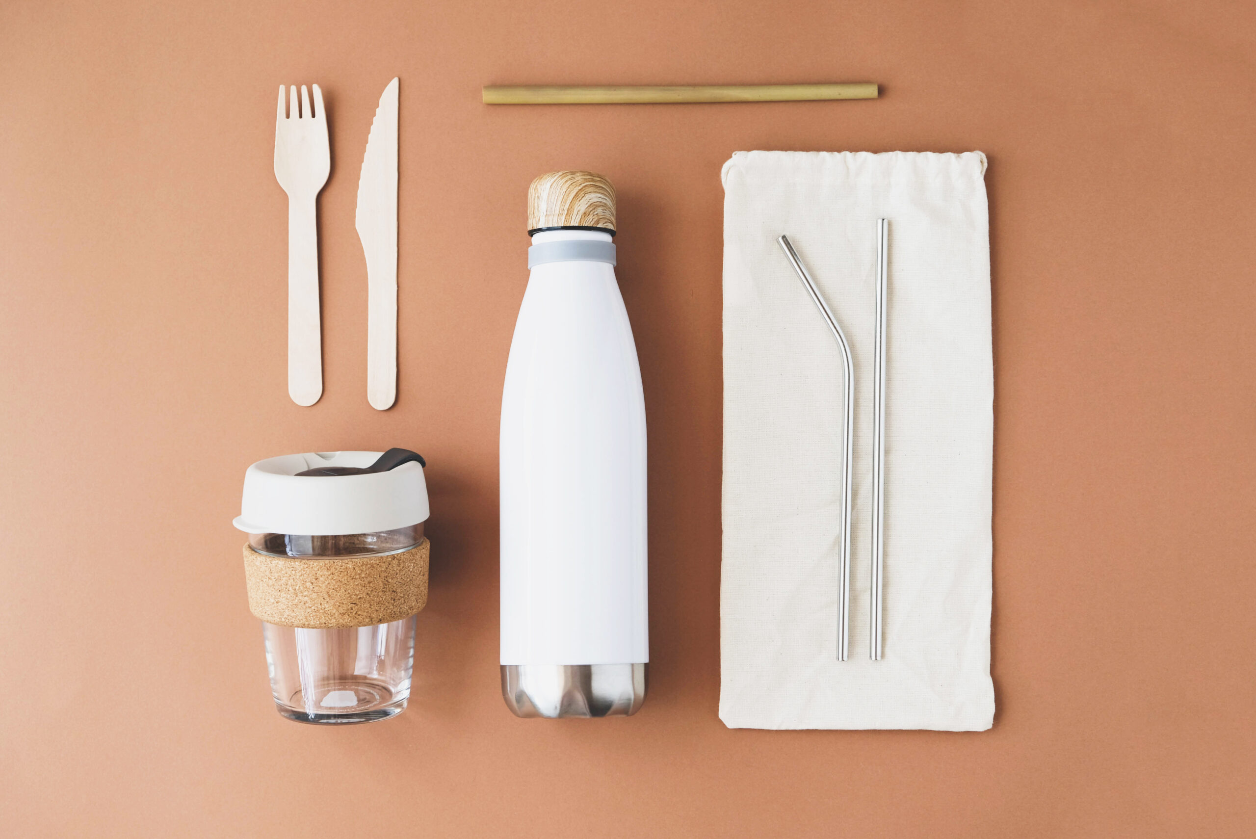 Stylish reusable eco friendly items on brown background. Metal straws and flask, bamboo straw, glass coffee cup, wooden cutlery. Zero waste, plastic free, sustainable lifestyle concept.